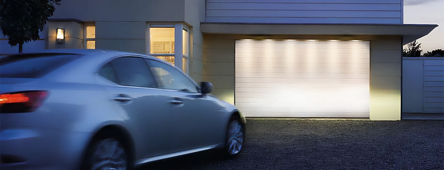 Moderne Hausfassade bei Abenddämmerung mit beleuchtetem, geschlossenem Garagentor und vorbeifahrendem Pkw in der Einfahrt.