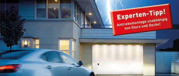 Moderne Hausfassade bei Abenddämmerung mit beleuchtetem, geschlossenem Garagentor und vorbeifahrendem Pkw in der Einfahrt.